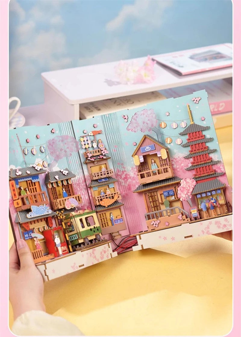 DIY book nook kit “Sakura Market,” featuring a glowing cherry blossom-lined miniature street scene with lanterns, shops, umbrellas, and Japanese architecture. Ideal for DIY and crafts lovers and fans of tiny dollhouses and book nook displays.