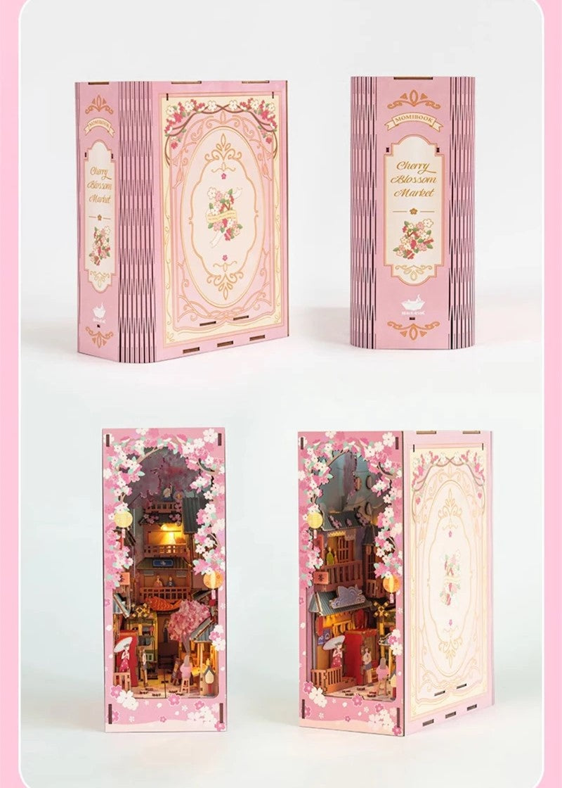 DIY book nook kit “Sakura Market,” featuring a glowing cherry blossom-lined miniature street scene with lanterns, shops, umbrellas, and Japanese architecture. Ideal for DIY and crafts lovers and fans of tiny dollhouses and book nook displays.
