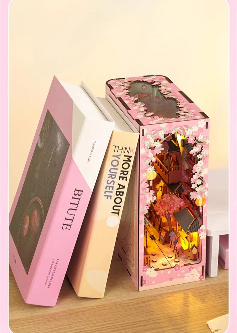 DIY book nook kit “Sakura Market,” featuring a glowing cherry blossom-lined miniature street scene with lanterns, shops, umbrellas, and Japanese architecture. Ideal for DIY and crafts lovers and fans of tiny dollhouses and book nook displays.