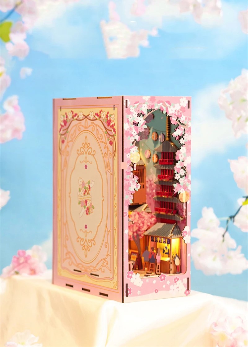 DIY book nook kit “Sakura Market,” featuring a glowing cherry blossom-lined miniature street scene with lanterns, shops, umbrellas, and Japanese architecture. Ideal for DIY and crafts lovers and fans of tiny dollhouses and book nook displays.