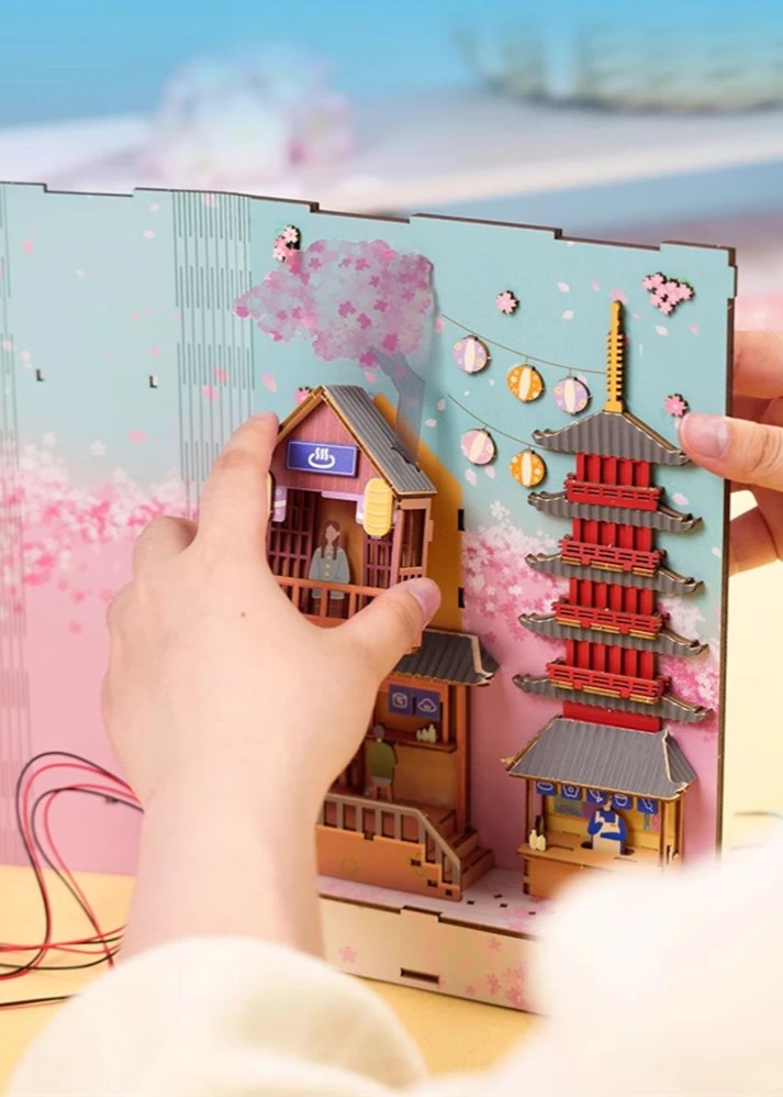 DIY book nook kit “Sakura Market,” featuring a glowing cherry blossom-lined miniature street scene with lanterns, shops, umbrellas, and Japanese architecture. Ideal for DIY and crafts lovers and fans of tiny dollhouses and book nook displays.
