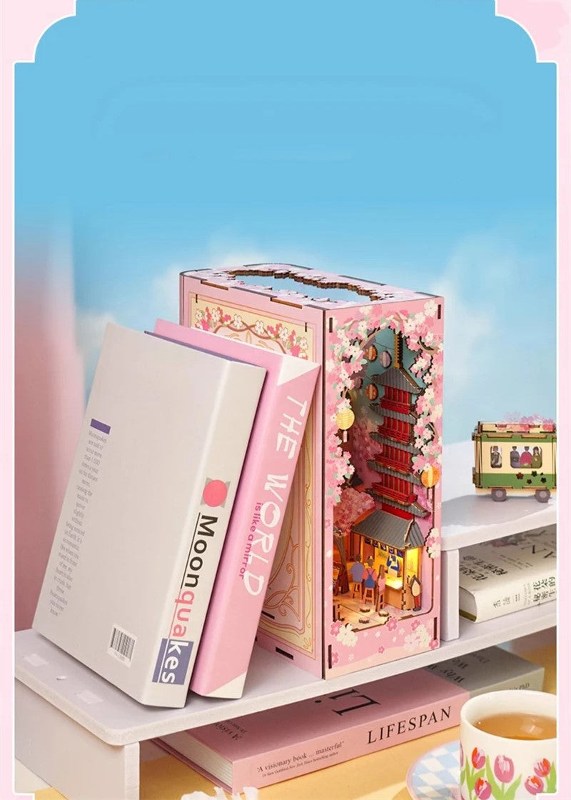 DIY book nook kit “Sakura Market,” featuring a glowing cherry blossom-lined miniature street scene with lanterns, shops, umbrellas, and Japanese architecture. Ideal for DIY and crafts lovers and fans of tiny dollhouses and book nook displays.