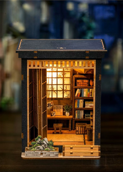 DIY book nook of a Japanese-style tea alcove featuring a glowing paper lantern, bonsai rock garden, shoji screen doors, bookshelves, and a peaceful wooden table, evoking the quiet ambiance of a neighborhood tea house.