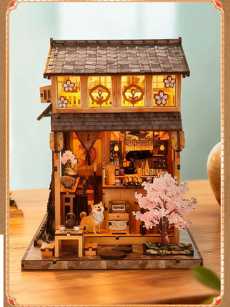 DIY miniature Japanese izakaya house featuring a cat chef, cherry blossom trees, hanging lanterns, a friendly Shiba Inu dog, and a warm glowing interior with tiny food items and sake bottles.