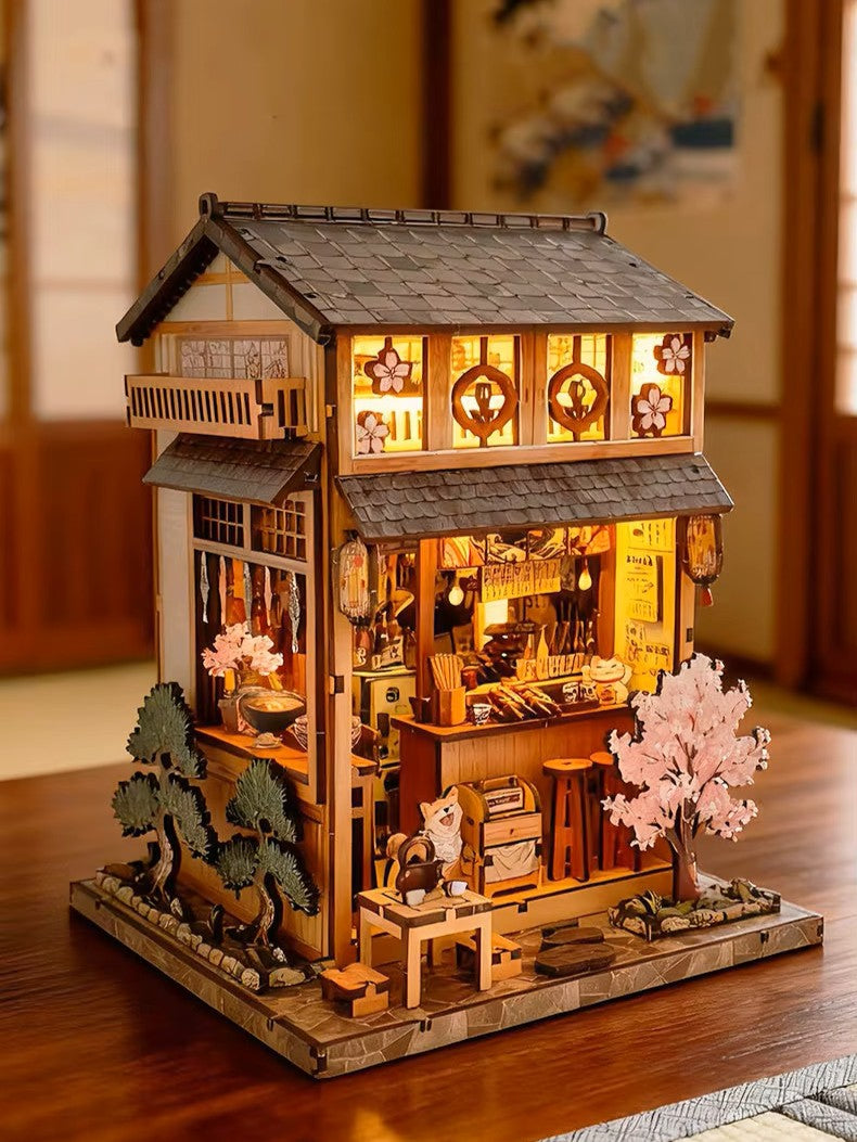 DIY miniature Japanese izakaya house featuring a cat chef, cherry blossom trees, hanging lanterns, a friendly Shiba Inu dog, and a warm glowing interior with tiny food items and sake bottles.