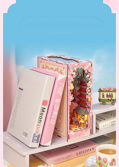 DIY book nook kit “Sakura Market,” featuring a glowing cherry blossom-lined miniature street scene with lanterns, shops, umbrellas, and Japanese architecture. Ideal for DIY and crafts lovers and fans of tiny dollhouses and book nook displays.
