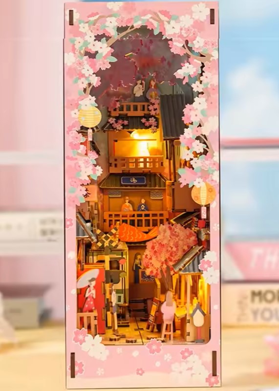 DIY book nook kit “Sakura Market,” featuring a glowing cherry blossom-lined miniature street scene with lanterns, shops, umbrellas, and Japanese architecture. Ideal for DIY and crafts lovers and fans of tiny dollhouses and book nook displays.
