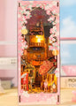 DIY book nook kit “Sakura Market,” featuring a glowing cherry blossom-lined miniature street scene with lanterns, shops, umbrellas, and Japanese architecture. Ideal for DIY and crafts lovers and fans of tiny dollhouses and book nook displays.