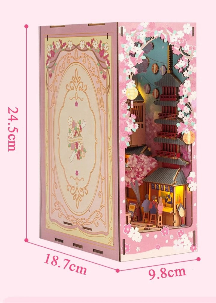 DIY book nook kit “Sakura Market,” featuring a glowing cherry blossom-lined miniature street scene with lanterns, shops, umbrellas, and Japanese architecture. Ideal for DIY and crafts lovers and fans of tiny dollhouses and book nook displays.
