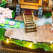 DIY miniature kit “Blossom Lantern Pavilion,” featuring a traditional East Asian-style building with curved roofs, cherry blossom trees, red lanterns, a waterfall, and glowing lights. A serene DIY and crafts project for fans of miniature homes and nature-inspired design.