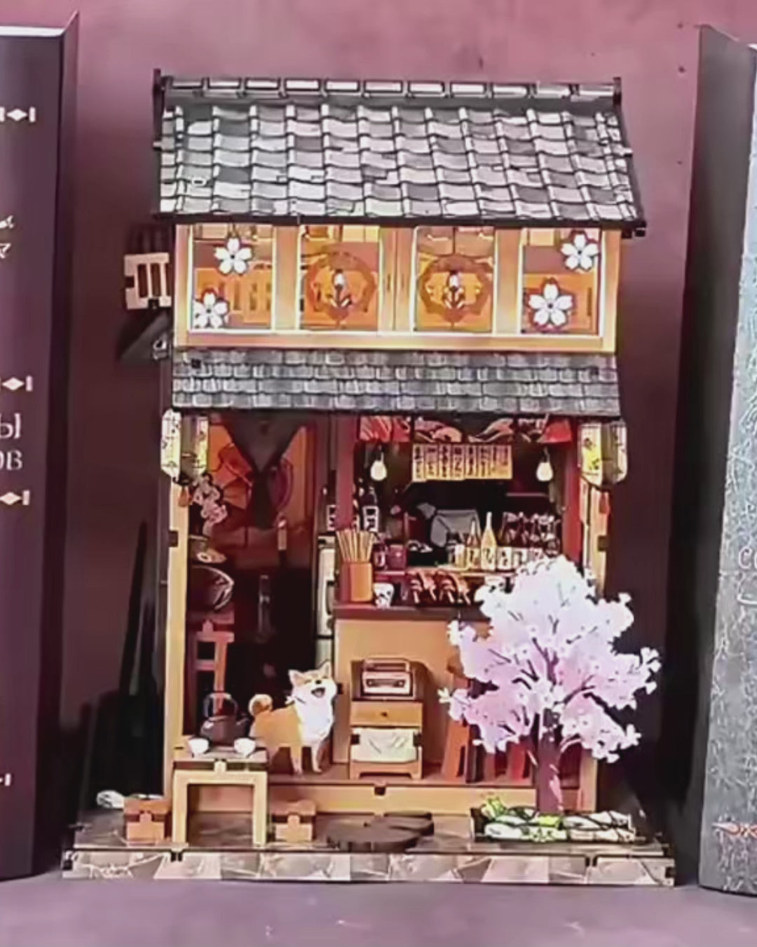 DIY miniature Japanese izakaya house featuring a cat chef, cherry blossom trees, hanging lanterns, a friendly Shiba Inu dog, and a warm glowing interior with tiny food items and sake bottles.