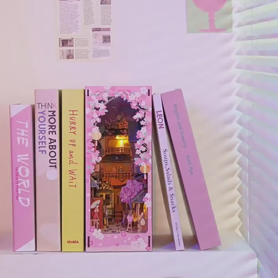DIY book nook kit “Sakura Market,” featuring a glowing cherry blossom-lined miniature street scene with lanterns, shops, umbrellas, and Japanese architecture. Ideal for DIY and crafts lovers and fans of tiny dollhouses and book nook displays.